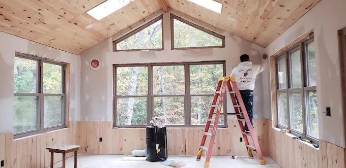 Worker on ladder painting interior of wooden cabin with large windows