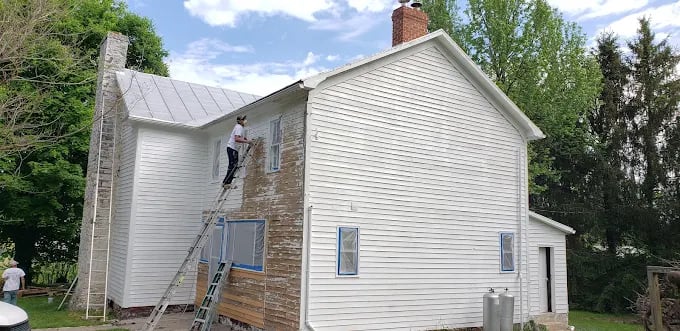Person painting white siding on two-story house using ladder on sunny day